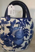 A ceramic handbag with a glossy white body decorated with blue floral and fruit motifs, featuring a dark blue handle and a hanging white tag.