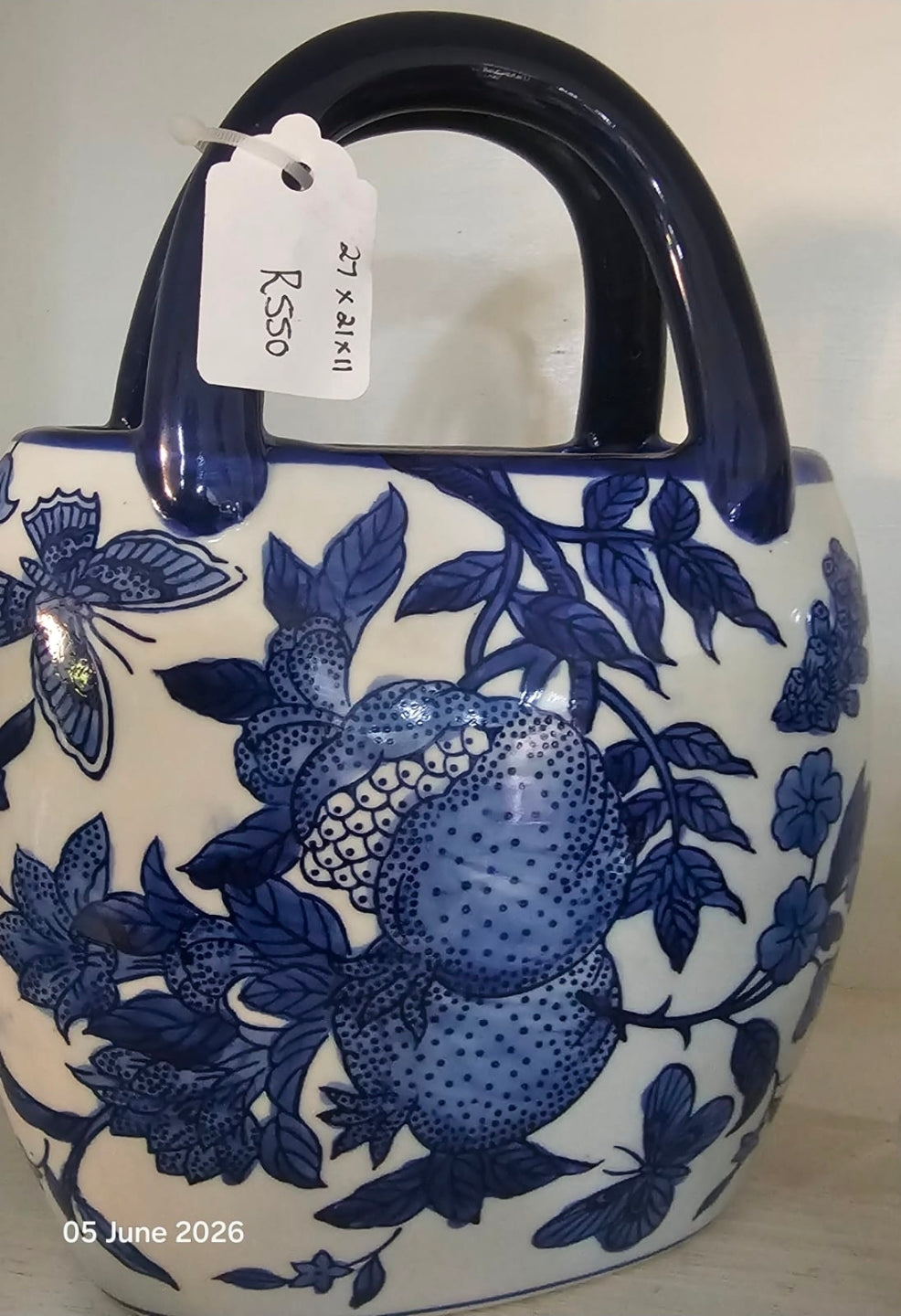 A ceramic handbag with a glossy white body decorated with blue floral and fruit motifs, featuring a dark blue handle and a hanging white tag.